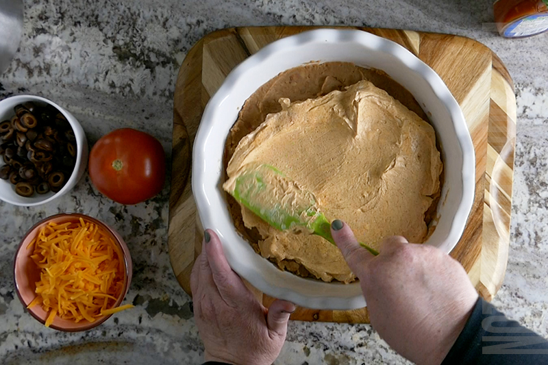 spreading the creamy taco mixture on top of the refried beans in the layered taco dip
