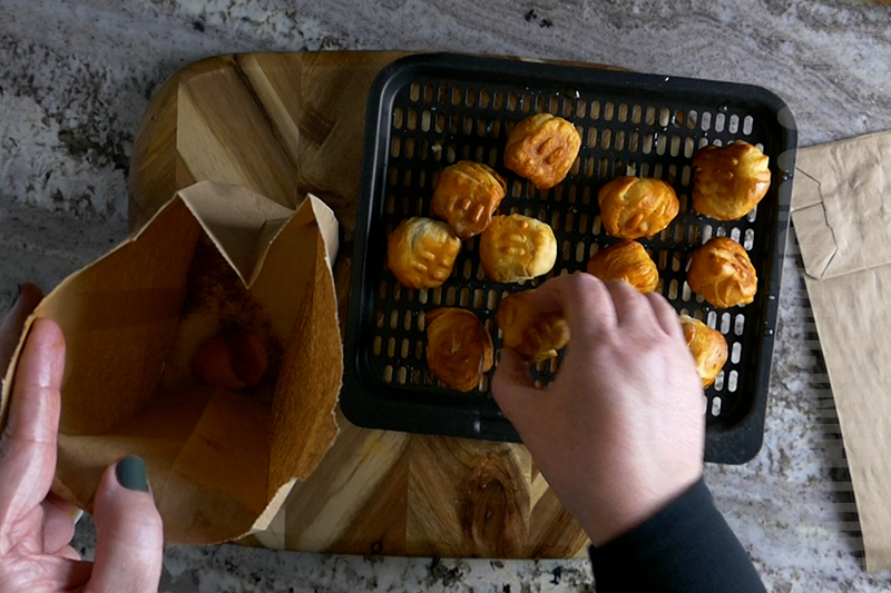 adding baked dough balls that have been brushed with melted butter to a paper lunch bag filled with sugar and cinnamon to coat the donuts