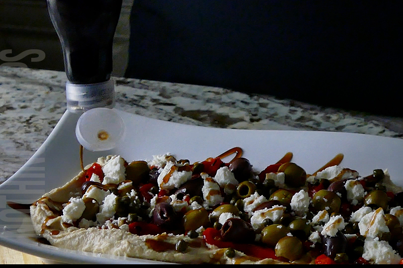drizzling balsamic glaze on hummus layered with roasted peppers, feta, capers and olives