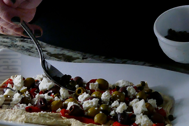 layering capers on hummus with roasted peppers and olives