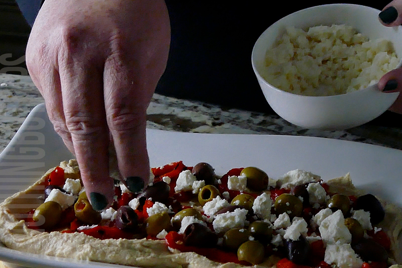 layering feta cheese on hummus with roasted peppers and olives
