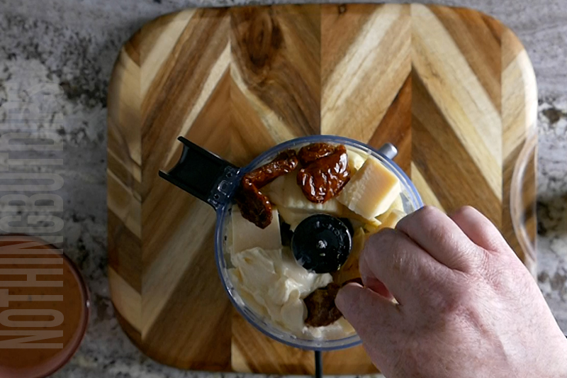 adding sun dried tomatoes to a mini food processor to make baked artichoke dip